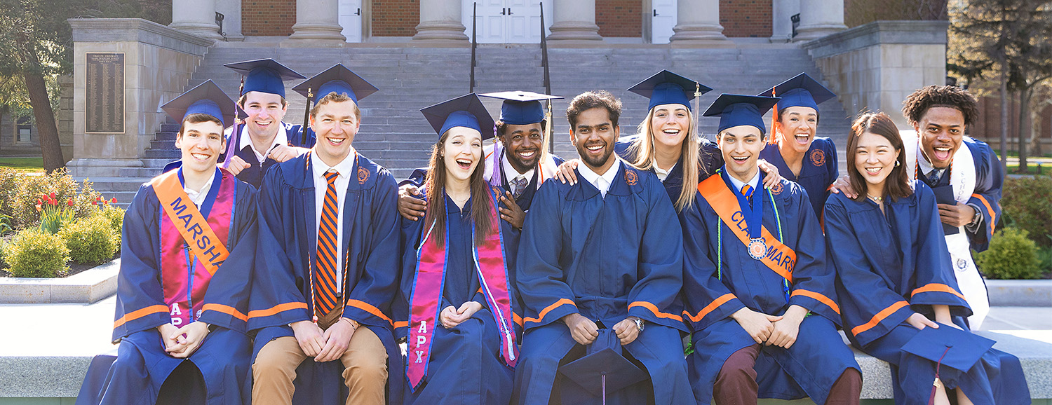 Group of graduates pictured on campus in front of Hendricks Chapel banner
				
