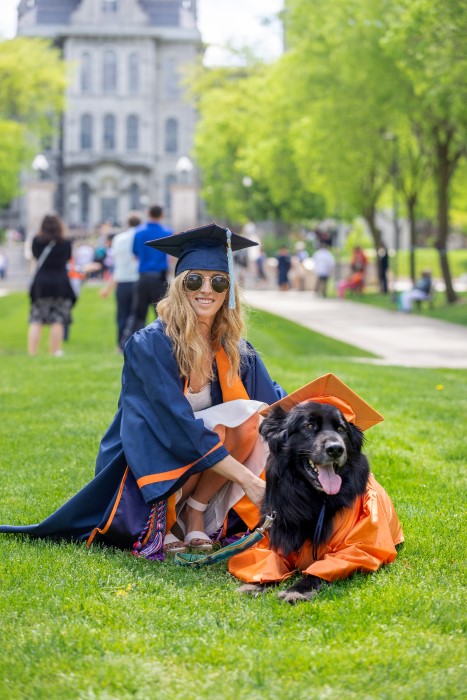 Class of 2022 graduate dressed in regalia poses with her dog – also dress in regalia – on campus in front of the Hall of Languages