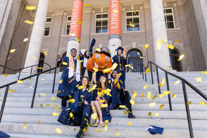 Group of graduates dressed in regalia celebrate with Otto on the stairs of Carnegie Library with confetti in the foreground